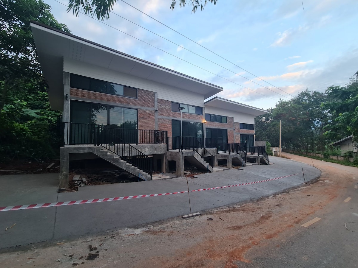 A modern building is shown with large glass windows and a combination of brick and concrete exterior. The structure features two levels with exterior staircases leading to the upper floors. Surrounding greenery and a paved road create a serene environment.