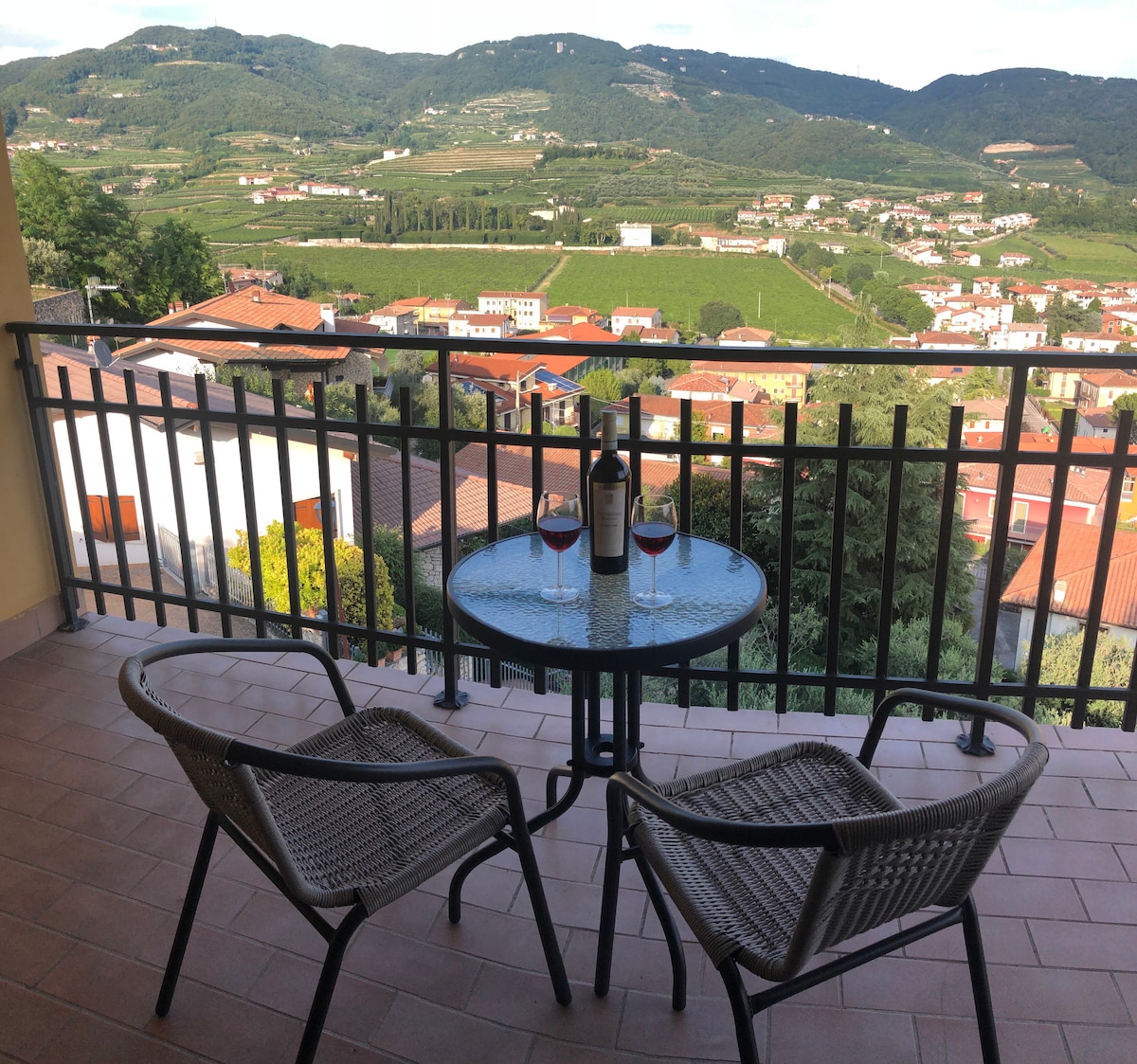 An inviting terrace features a glass table with two chairs, overlooking a picturesque landscape of rolling hills and rooftops. A bottle of wine and two glasses are placed on the table, enhancing the serene outdoor experience.