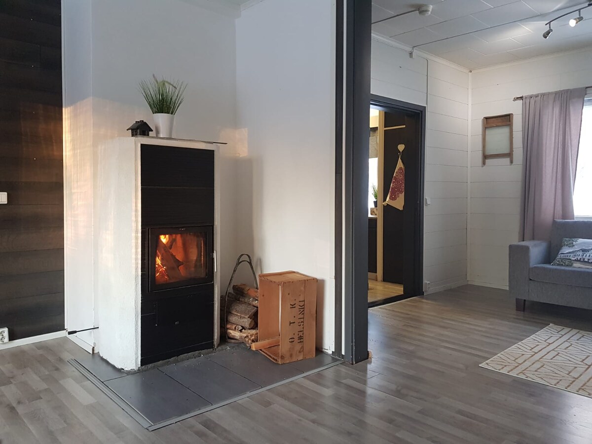 A warm living area features a modern fireplace, with flickering flames visible through a glass front. A cozy couch is positioned nearby, accompanied by a small storage box for firewood. Natural light filters in through a window, softened by light curtains.