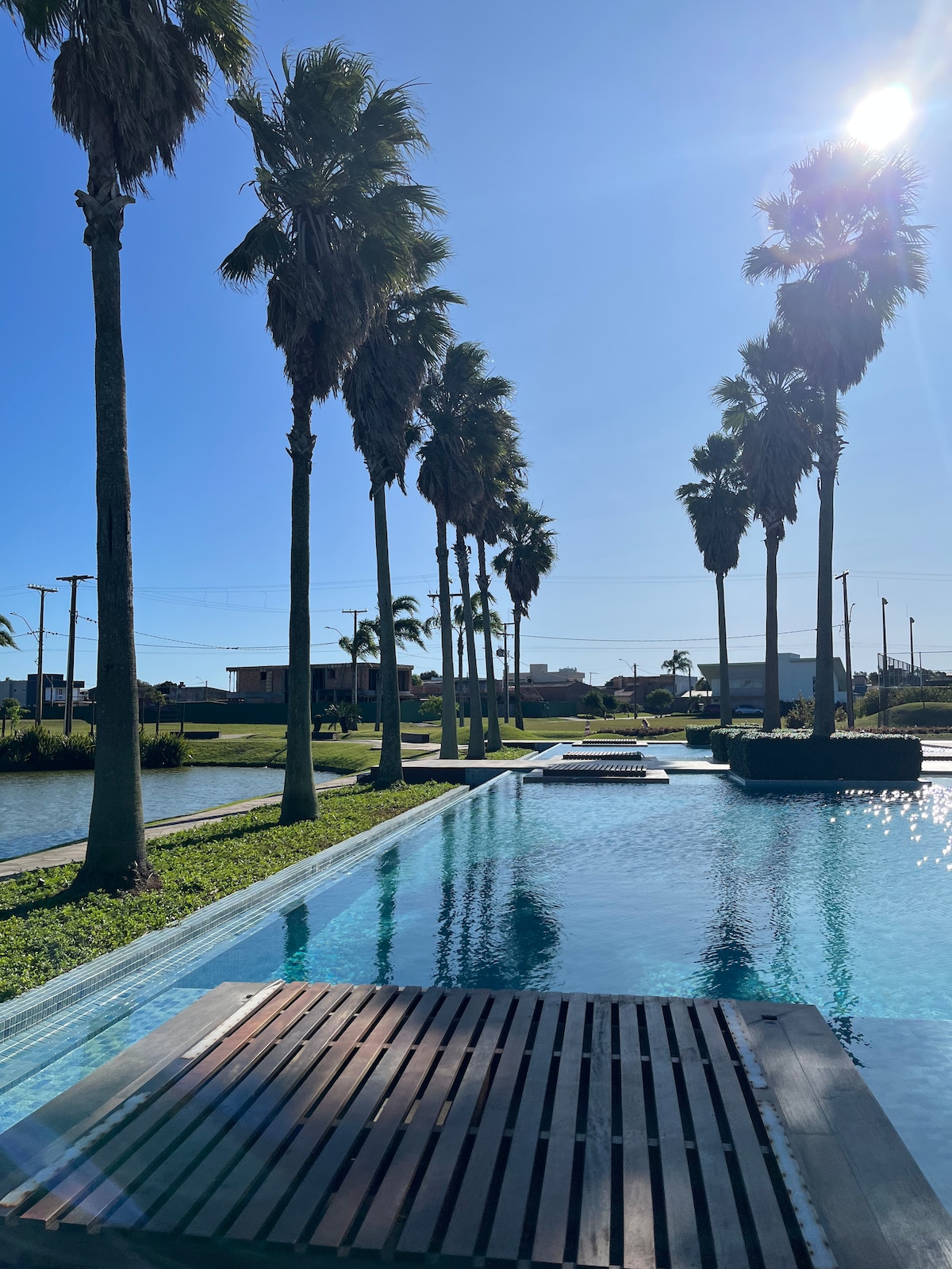 The image displays a serene pool area lined with tall palm trees that gently sway in the breeze. Wooden deck platforms are positioned within the water, providing a space for relaxation. Clear blue skies reflect off the tranquil surface of the pool, enhancing the outdoor setting.