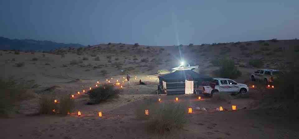 A desert scene is captured at twilight, featuring a central tent surrounded by softly glowing lanterns on the sand. Vehicles are parked nearby, and distant hills are faintly visible under the evening sky, creating a serene atmosphere.