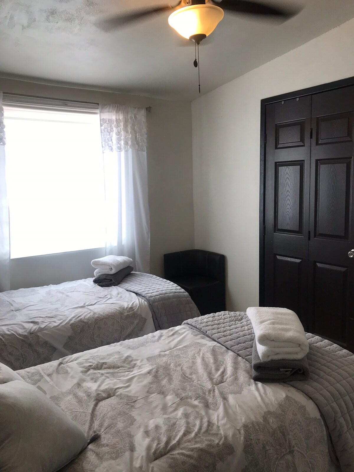 A bright bedroom features two twin beds dressed in light-colored linens with neatly arranged towels. A window with sheer curtains allows natural light to fill the room. A corner shelf unit and a dark wooden door complete the warm and functional space.
