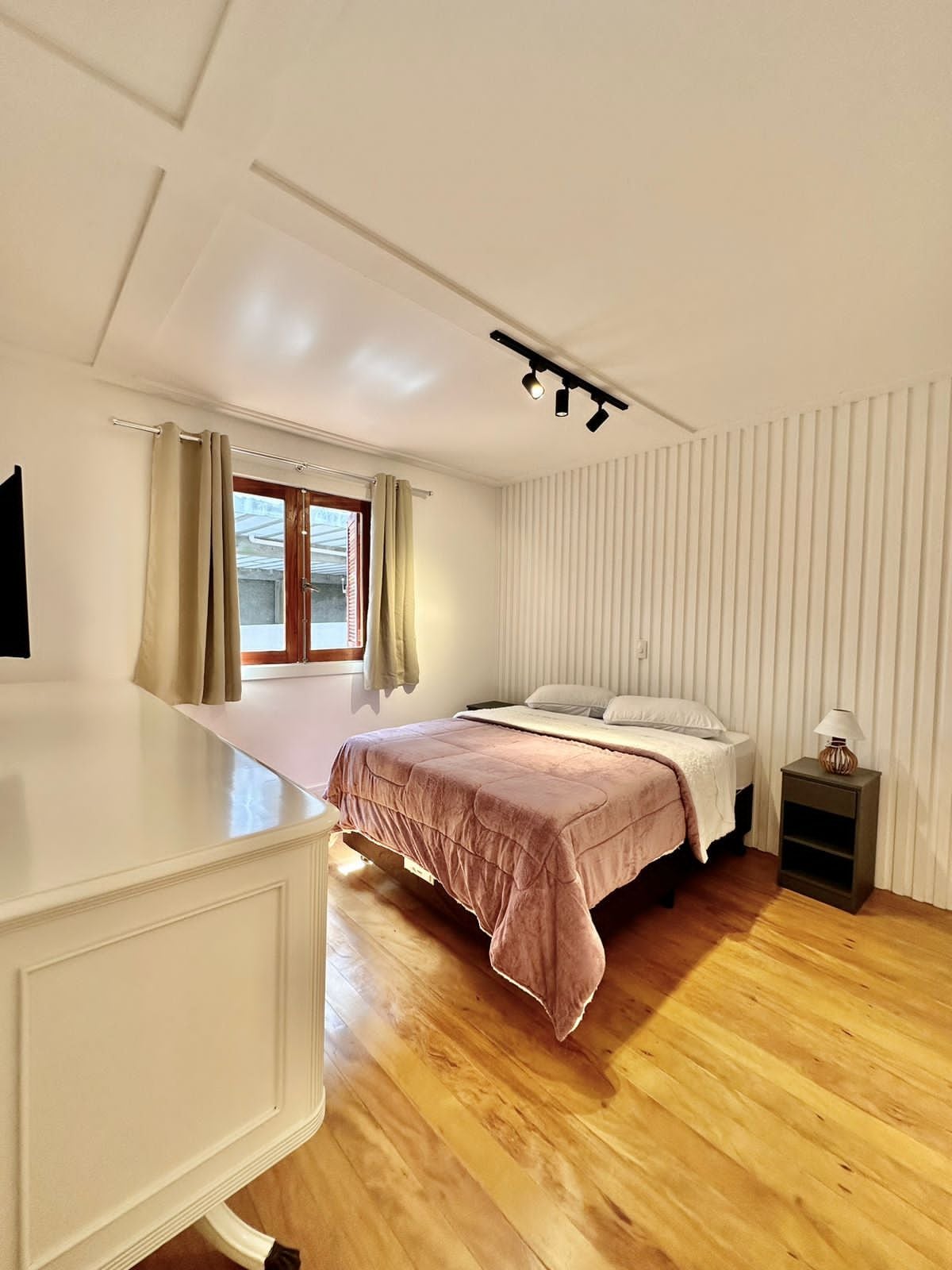 A well-lit bedroom features a spacious bed with a soft pink blanket. Natural light floods in through a window adorned with light curtains. The room is accented by wooden flooring and a subtle striped wall design. A nightstand and small lamp add to the decor.