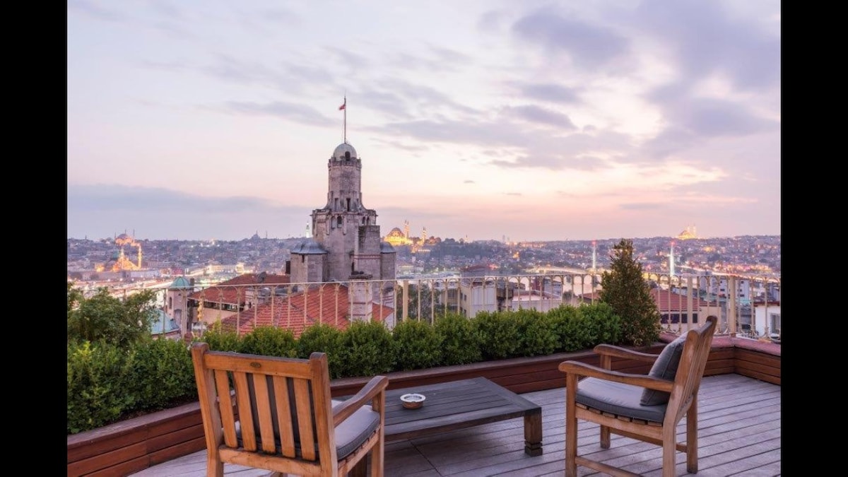 A large rooftop terrace features wooden seating with a coffee table, overlooking a panoramic view of the city. The silhouette of Galata Tower is visible against the skyline, while greenery lines the terrace perimeter. Soft colors of dusk enhance the tranquil setting.