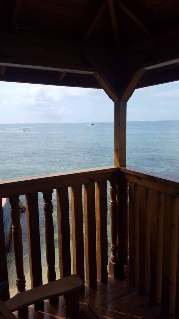 The image captures a wooden balcony overlooking the calm Caribbean Sea, with a clear view of the water stretching to the horizon. Small boats can be seen gently bobbing on the surface, under a partly cloudy sky, enhancing the serene coastal atmosphere.