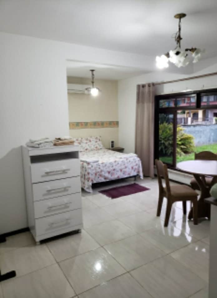 A clean and bright room features a floral-patterned bedspread draped over a spacious bed. A window allows natural light to enter, while a small dining table with chairs is positioned nearby. A dresser stands in the corner, enhancing the room's functionality.