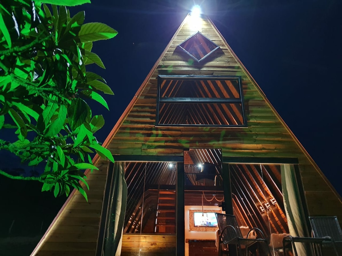A charming A-frame structure features a wooden exterior with large windows, illuminating the interior from soft lighting. The triangular roof enhances the upward space while greenery is visible in the foreground, contributing to the natural setting.