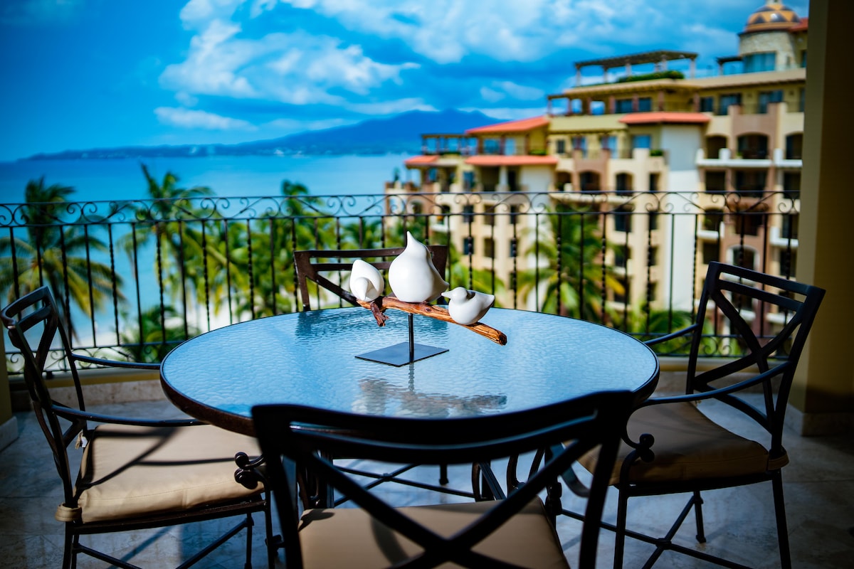 A round table with a glass top is set against a scenic view, displaying a decorative piece of three white birds atop a branch. Black chairs with cushioned seats surround the table, while the backdrop includes lush palm trees and a distant ocean, under a bright blue sky.