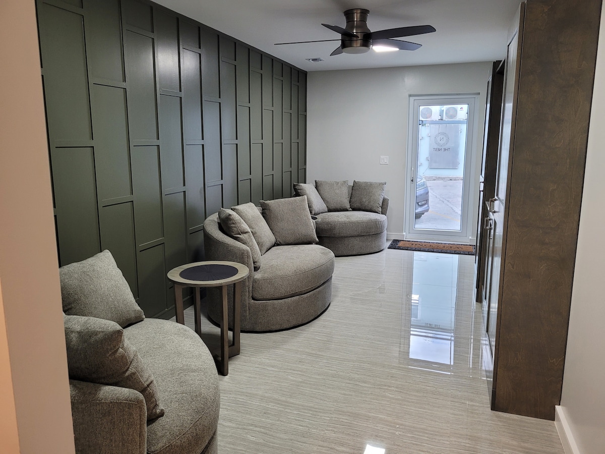 A modern sitting area features a curved sectional sofa and matching armchairs, arranged around a small round table. A sleek ceiling fan is installed above, enhancing airflow. Natural light enters through a glass door, which leads to an outdoor space.