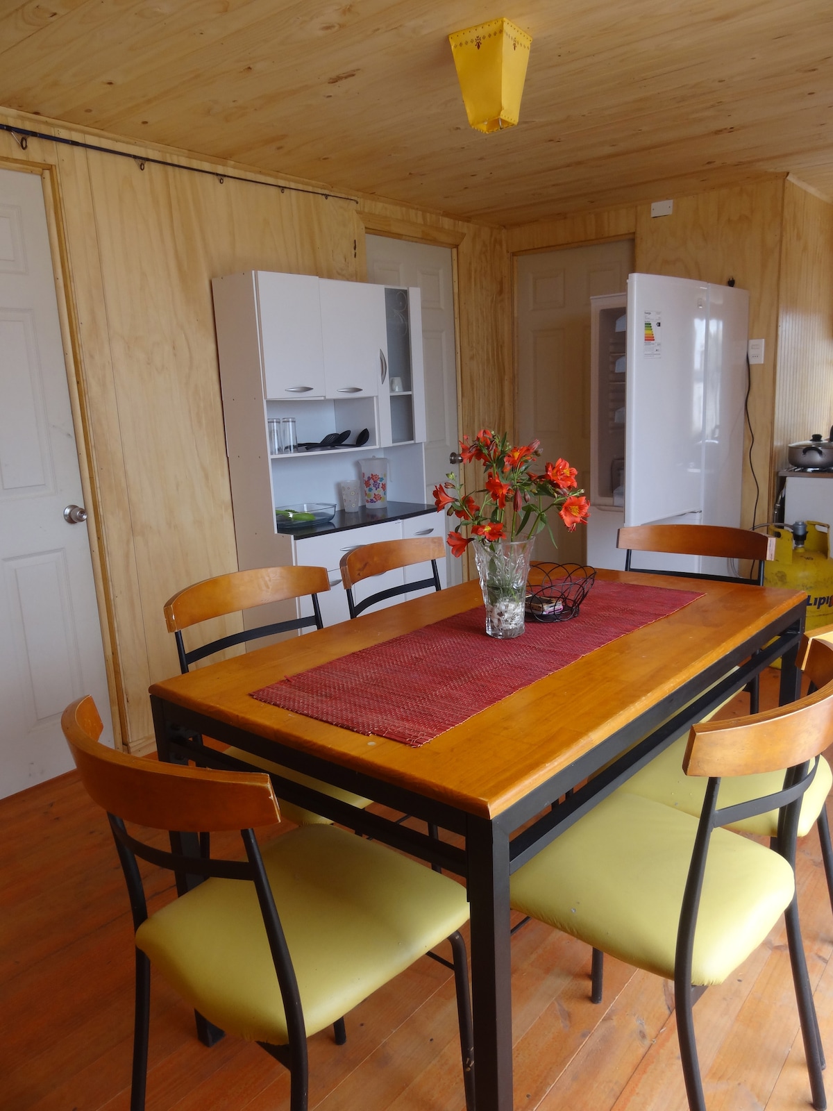 A dining area is presented with a wooden table surrounded by six chairs. A vase filled with flowers is centrally placed on the table. A white cabinet and refrigerator are visible in the background, complemented by a light wooden wall and warm wooden flooring.