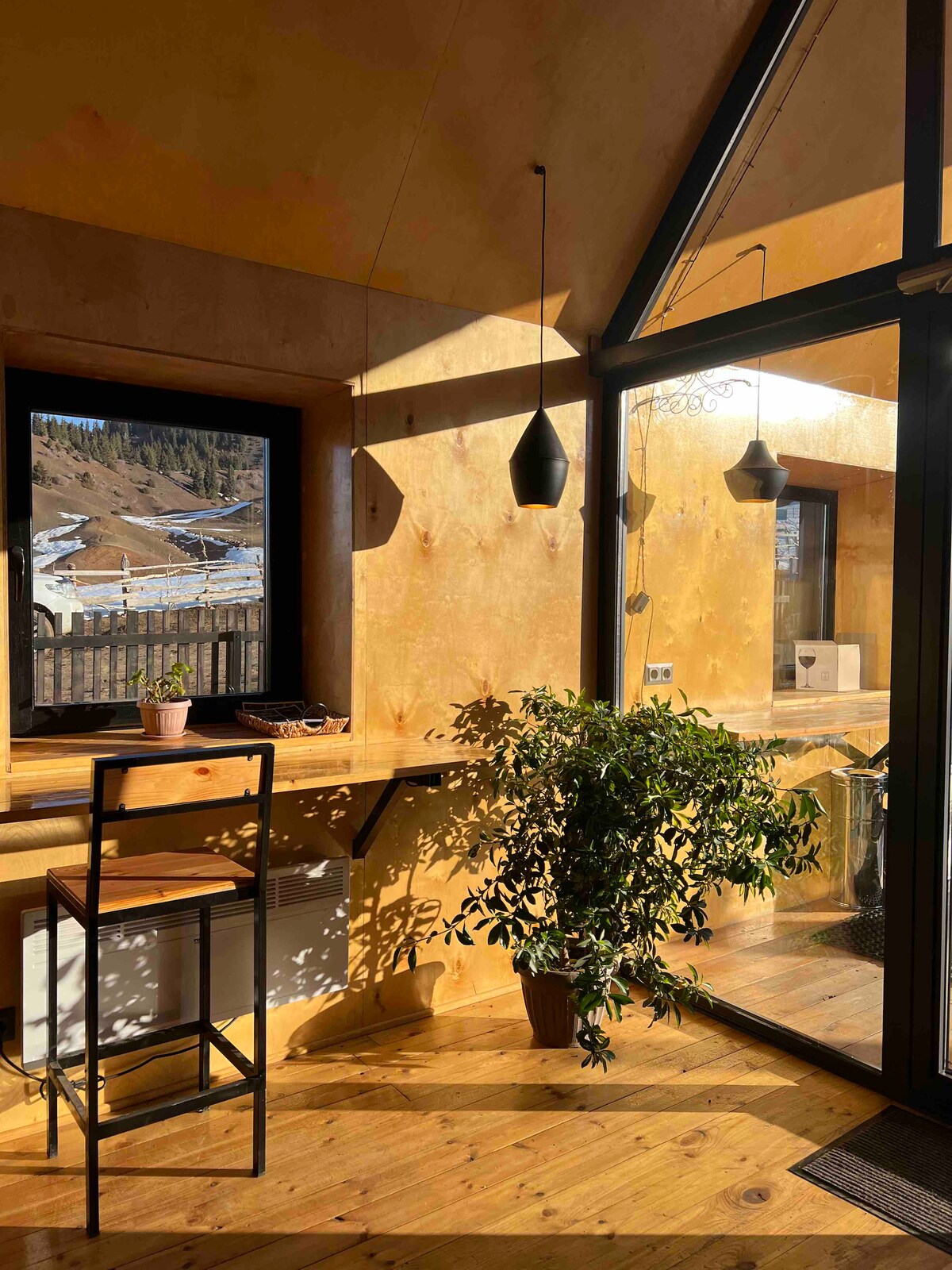 A sunlit corner features a wooden bar with stools beneath pendant lighting. Greenery on the counter complements the natural wood accents, while large windows provide views of the surrounding mountainous landscape.