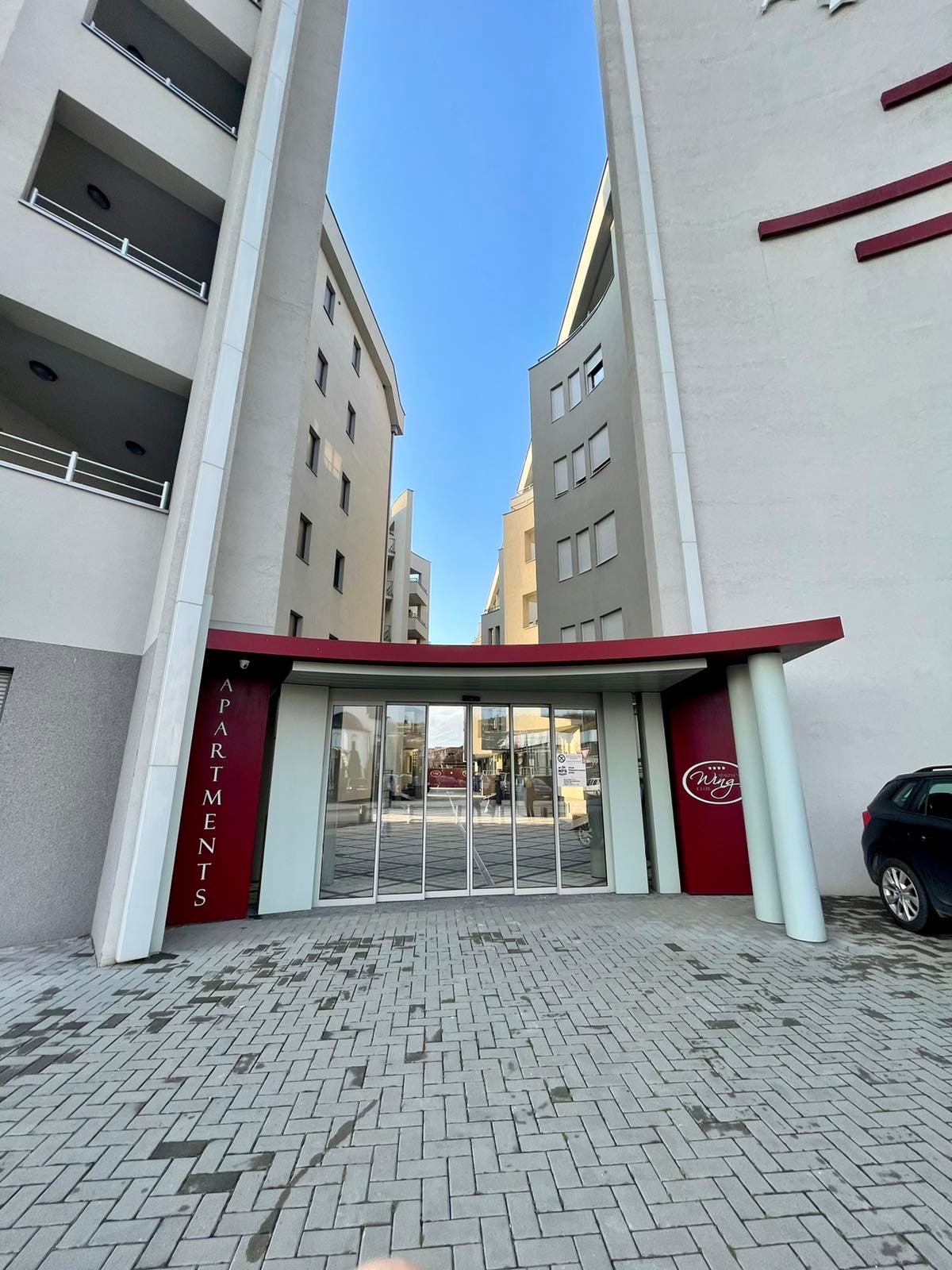 A modern building entrance is framed by two adjacent structures, featuring large glass doors marked with 'APARTMENTS.' The entrance is surrounded by a paved area and is bathed in natural light under a clear blue sky.