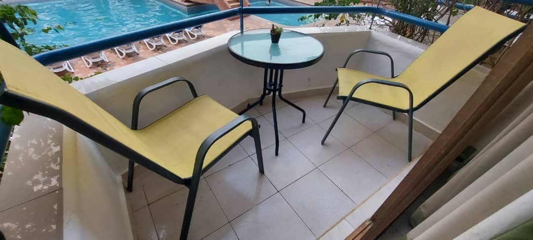A small balcony is furnished with two yellow loungers and a round table. A plant centerpiece rests on the table, and the view includes a swimming pool visible in the background. Potted greenery adds a touch of nature to the space.