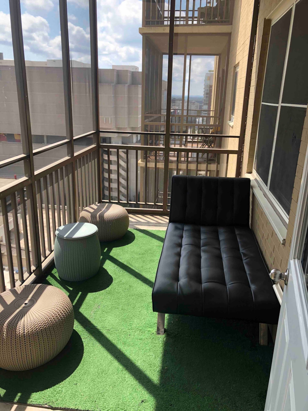 A covered balcony features a black chaise lounge and two textured round stools. The green artificial grass adds a touch of color, while large windows allow natural light to fill the space. The backdrop offers a glimpse of the surrounding cityscape.