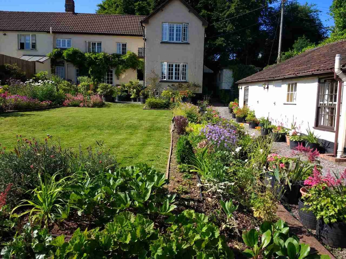 A charming property is accented by a well-maintained garden filled with a variety of colorful flowers and plants. Green lawn stretches across the foreground, leading to the inviting exterior of the house, which features multiple windows and a welcoming entryway surrounded by greenery.
