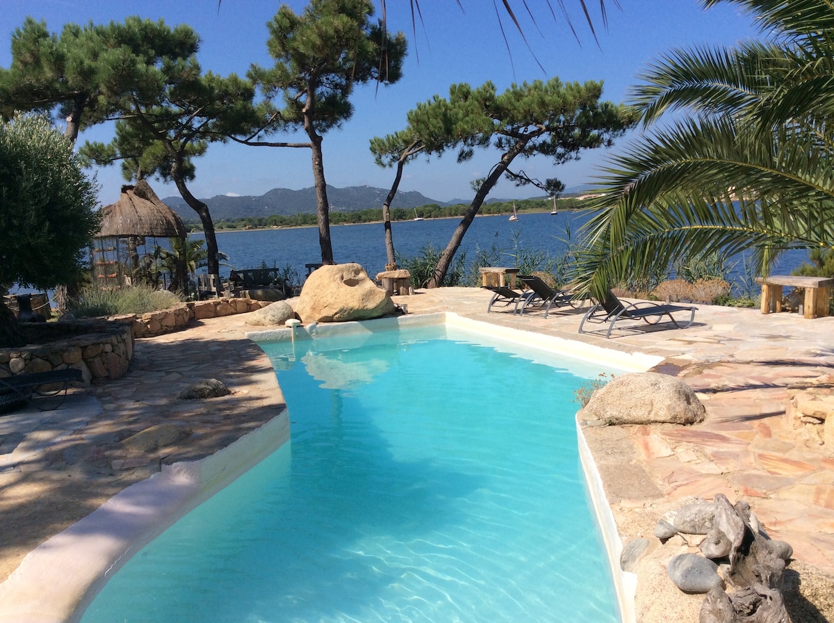 A tranquil pool area is surrounded by natural stone walls and lush vegetation. Comfortable lounge chairs are positioned on the deck, offering a serene view of the water. A thatched gazebo is visible in the distance, framed by swaying trees under a clear blue sky.