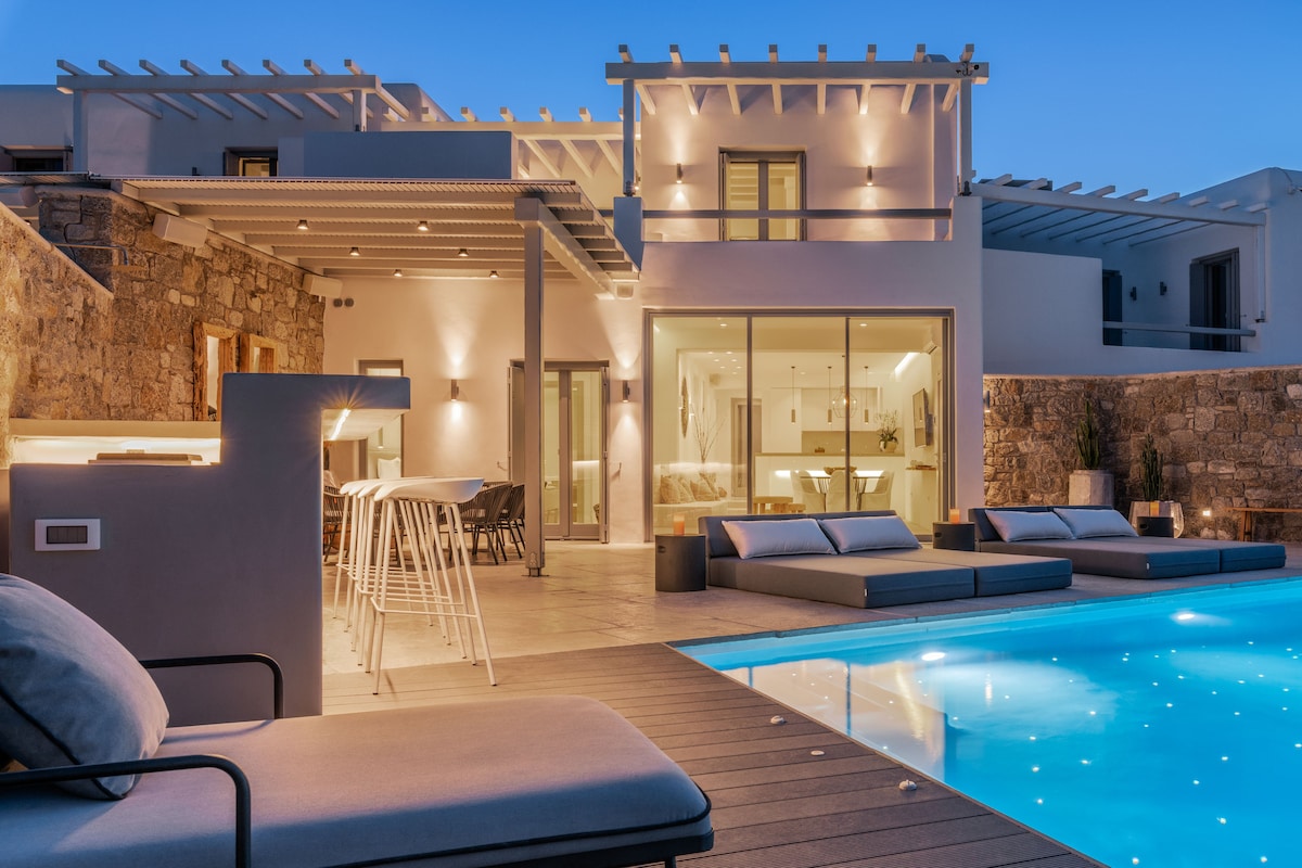 An outdoor space featuring a sparkling pool surrounded by stylish lounge chairs and a modern outdoor bar area. The villa's architecture showcases large glass doors and soft lighting, creating a welcoming atmosphere as evening sets in.