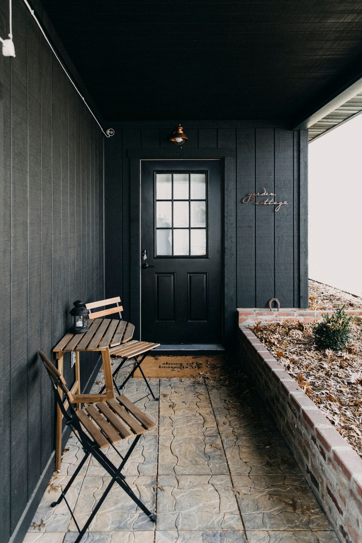 A cozy entrance features a black door framed by a decorative light. Two wooden folding chairs and a small table sit on a patterned stone surface, complemented by a small landscaped area with shrubs. A welcome mat adds a warm touch.
