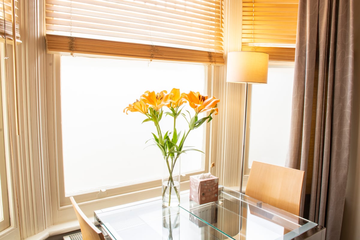 A glass dining table is positioned near a window, adorned with a vase of fresh orange lilies. Soft light filters through wooden blinds, creating a warm ambiance. A lamp provides additional illumination, and a discreet tissue box is placed on the table.