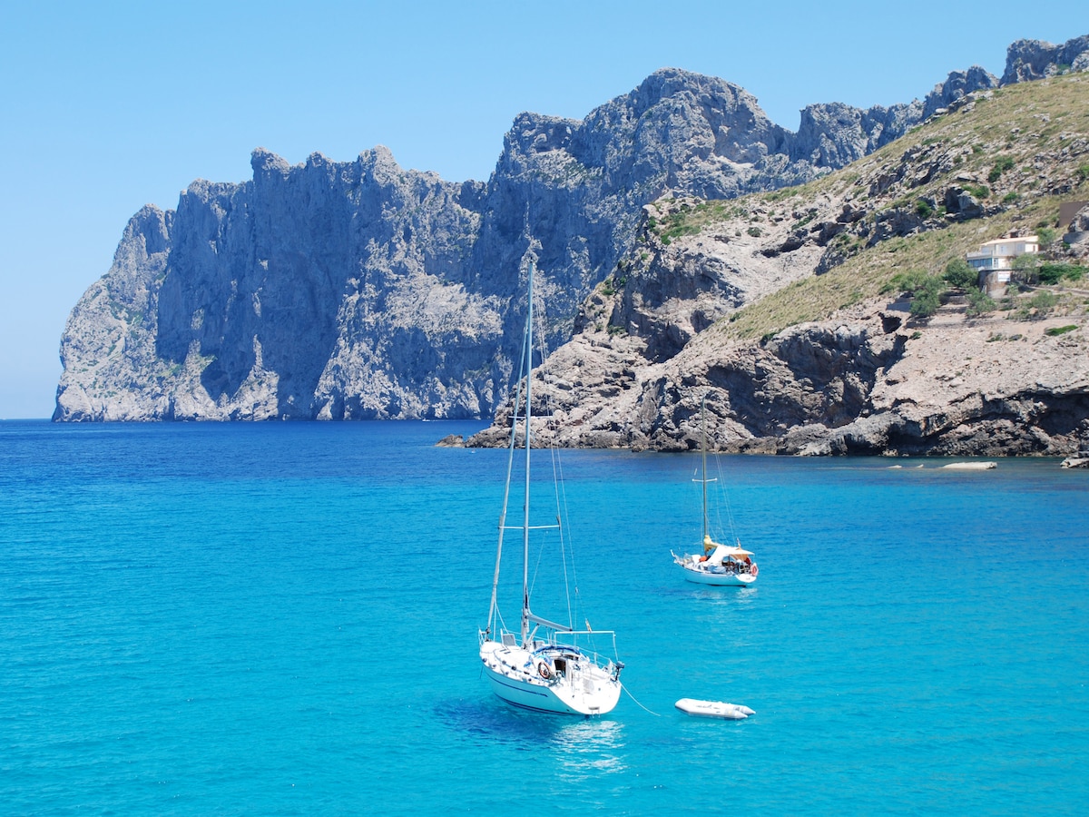 Two sailboats are anchored in crystal clear turquoise waters, surrounded by rugged cliffs that rise dramatically from the coastline. The inviting blue of the sea contrasts with the rocky landscape, creating a serene and picturesque coastal scene.