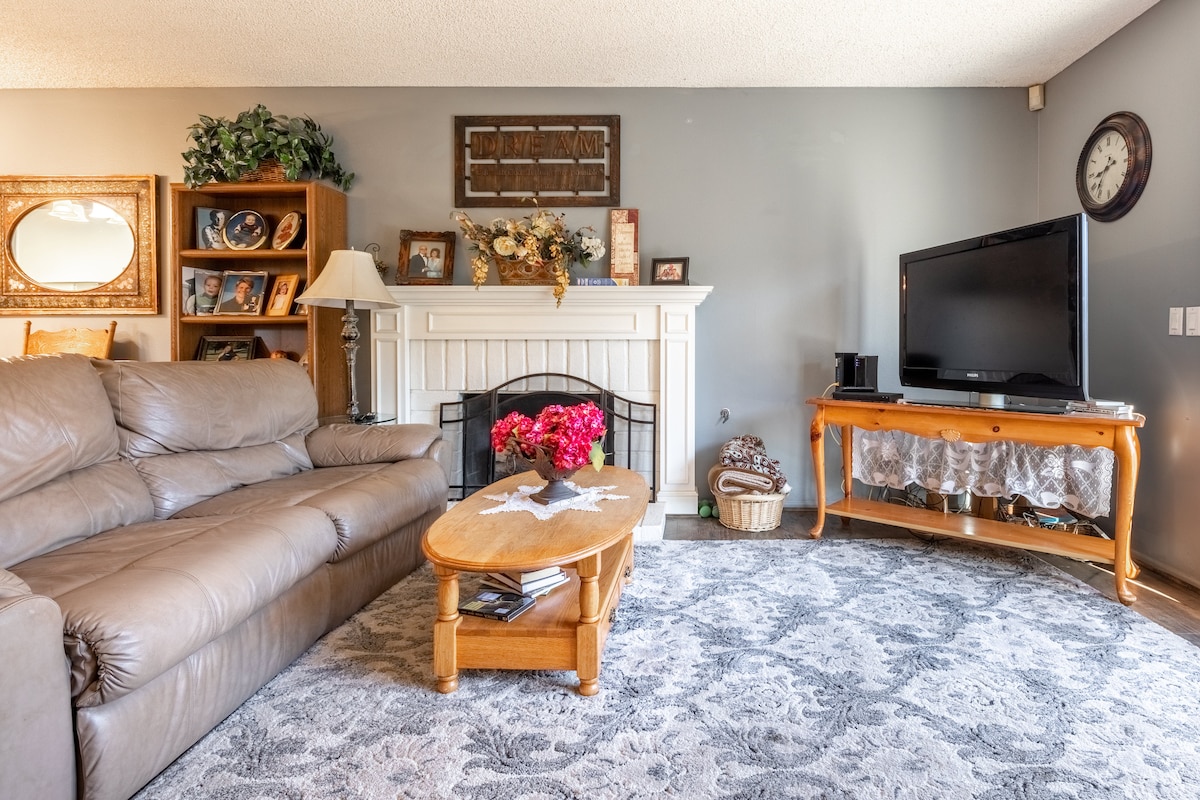 A cozy living room features a comfortable leather sofa and a wooden coffee table adorned with flowers. A decorative mirror and framed pictures enhance the decor, while a fireplace and a large television provide a focal point. A patterned area rug complements the space.