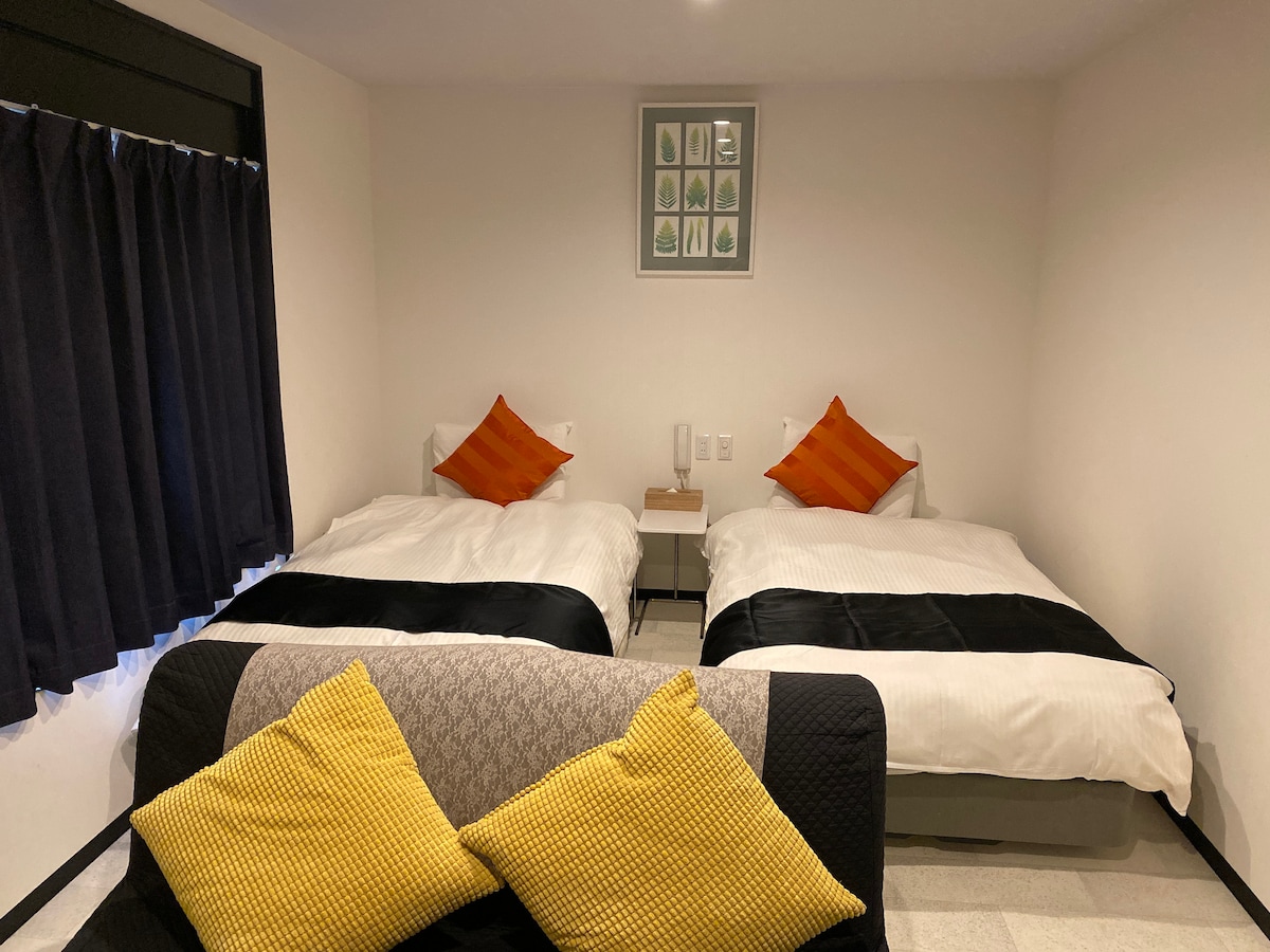The room features two spacious beds arranged with crisp white linens and contrasting black accents. Bright orange throw pillows add a pop of color. A small bedside table is positioned between the beds. Dark curtains frame a window, creating a cozy atmosphere.