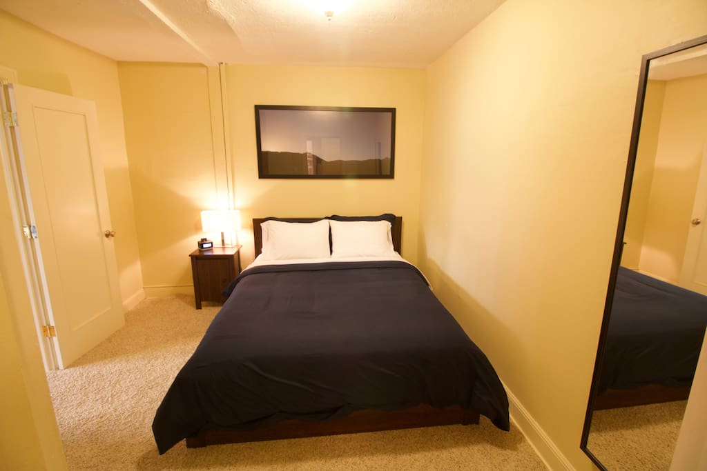 Bedroom features a full size wall mounted mirror, queen bed with luxury linens, closet with hangers and luggage rack, iPhone dock and reading light on a nightstand.