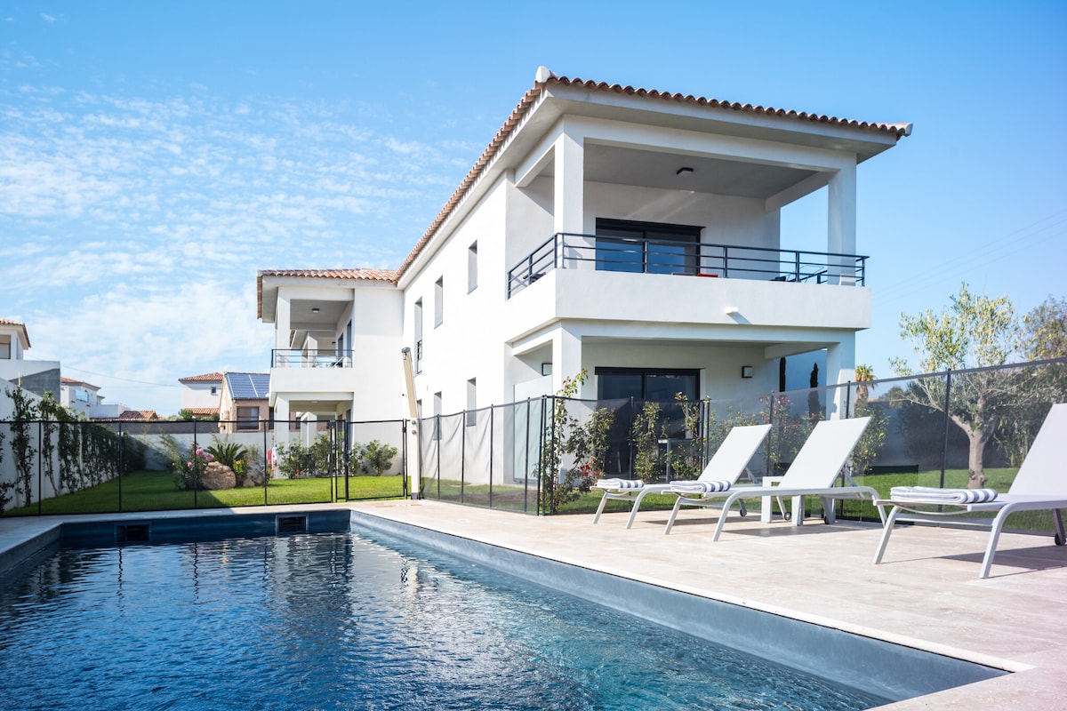 A modern two-story residence is seen with a refreshing outdoor pool in the foreground. The pool area includes lounge chairs arranged on a sun-drenched patio. Lush greenery and a fence enhance the outdoor space, while balconies offer views of the surrounding area.