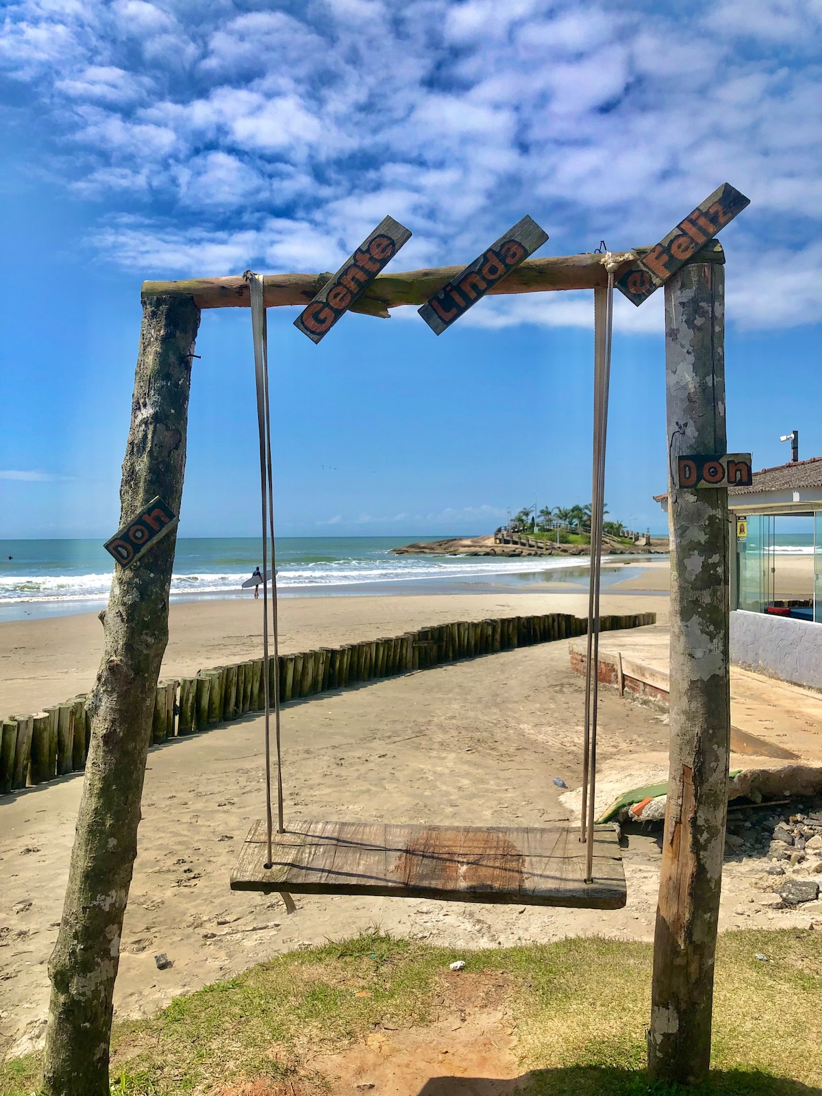 A swing made of wood is suspended between two tree trunks, with decorative signs attached. The swing faces a sandy beach with gentle waves lapping at the shore, and a clear blue sky overhead features scattered clouds.