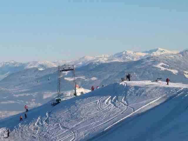 A snow-covered mountain landscape is displayed, showcasing ski slopes and a ski lift in the distance. Skiers can be seen navigating the slopes, while the expansive views of distant mountains and a clear blue sky provide a serene backdrop.