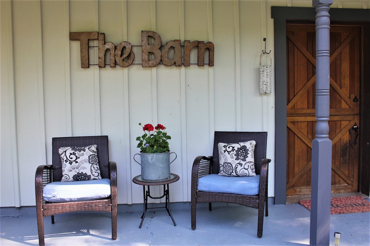 A welcoming entrance features two wicker chairs with soft cushions, positioned around a small table. A flower pot adds a touch of color, while a wooden sign displaying 'The Barn' is affixed to the exterior wall. A rustic door leads inside.