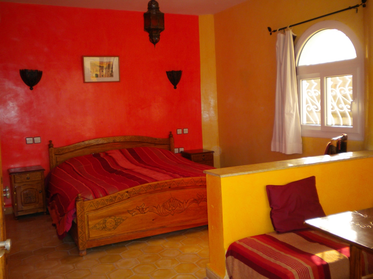 A warm-toned room features a spacious bed with rich red linens, complemented by a decorative wooden headboard. Walls are adorned in vibrant orange and red hues. A cozy seating area with a small table lies in the corner, and natural light enters through an elegantly arched window.