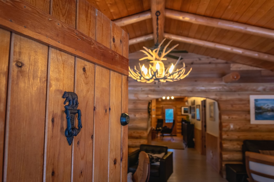 The entrance door features rustic wood with a bear-shaped knocker. The interior showcases warm wood paneling and a unique chandelier made of antlers, leading to a spacious living area visible in the background.