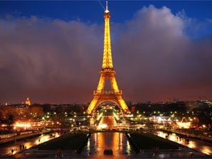 The Eiffel Tower is illuminated against a twilight sky, showcasing its intricate iron latticework. Surrounding areas are filled with lights from nearby buildings, while a reflective body of water enhances the scene's tranquility.