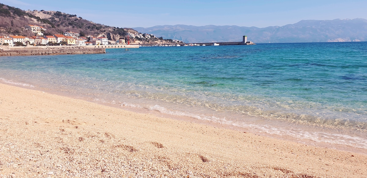 A serene beach scene is depicted, featuring golden sand meeting clear turquoise waters. Gentle waves lap the shore, while a distant coastline with colorful buildings is softly visible against the backdrop of rolling hills and a blue sky.
