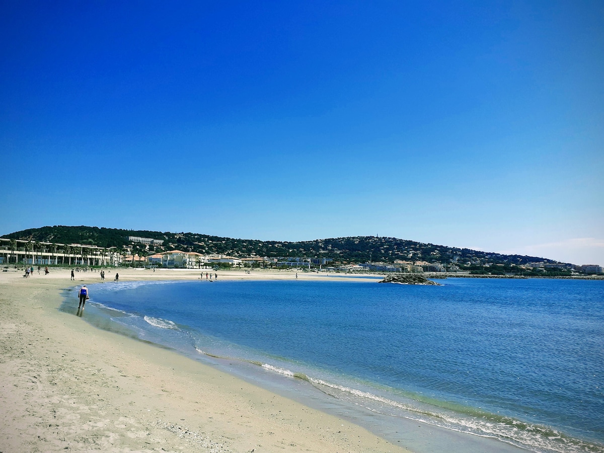 A scenic beach scene features soft sand leading to calm blue waters. Gentle waves lap at the shore, while distant hills provide a backdrop under a clear, blue sky. People stroll along the beach, enjoying the serene environment.