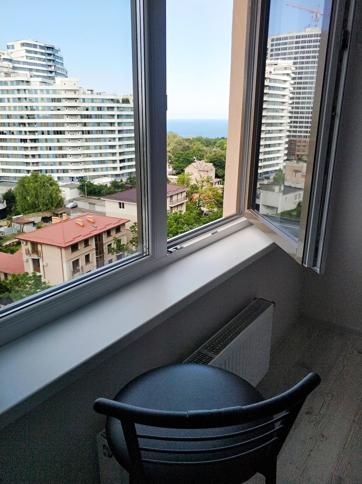 A window is open, offering a view of the sea and nearby buildings. A black chair is positioned by the window, providing a spot to enjoy the scenery. The space is filled with natural light, enhancing the calm atmosphere of the room.