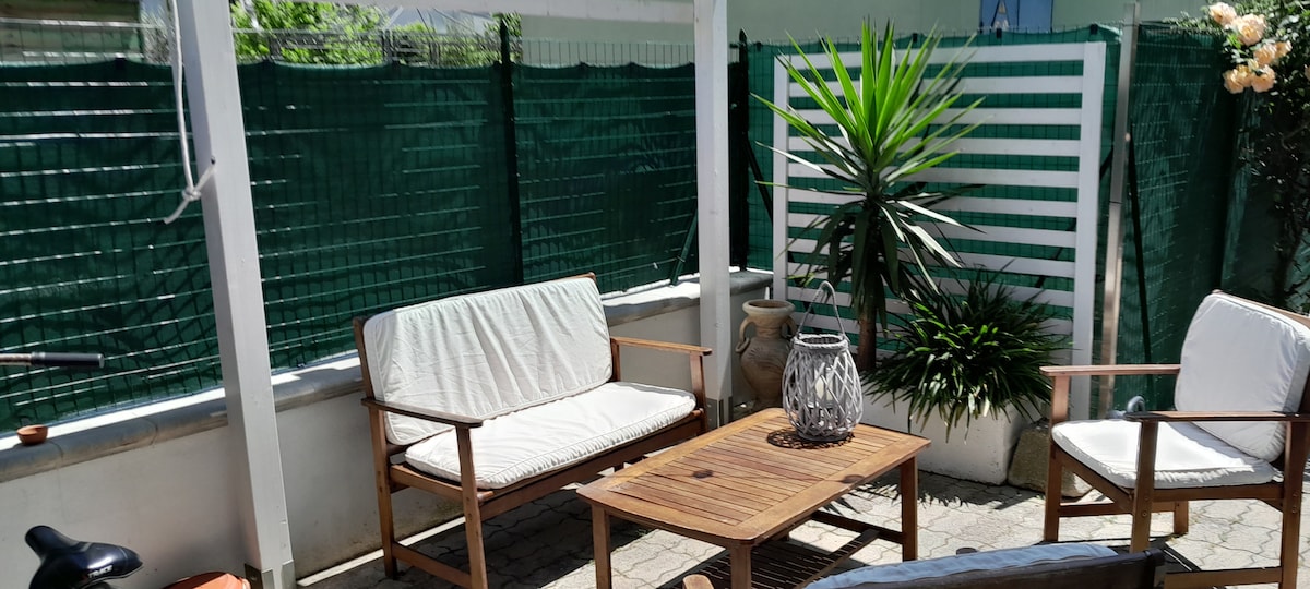 An outdoor seating area features two wooden chairs with light cushions and a matching wooden table. A decorative plant adds greenery, while a textured vase enhances the space. Lattice fencing provides privacy, with sunlight filtering through the structure above.
