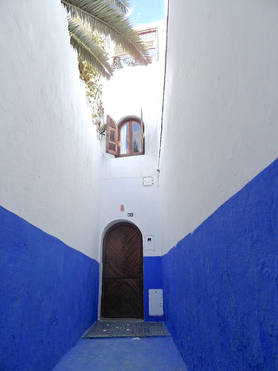 A narrow, sunlit alleyway is framed by whitewashed walls adorned with contrasting blue accents. A wooden door is set within the archway, above which a window with ornate shutters is partially open, allowing natural light to filter into the space.