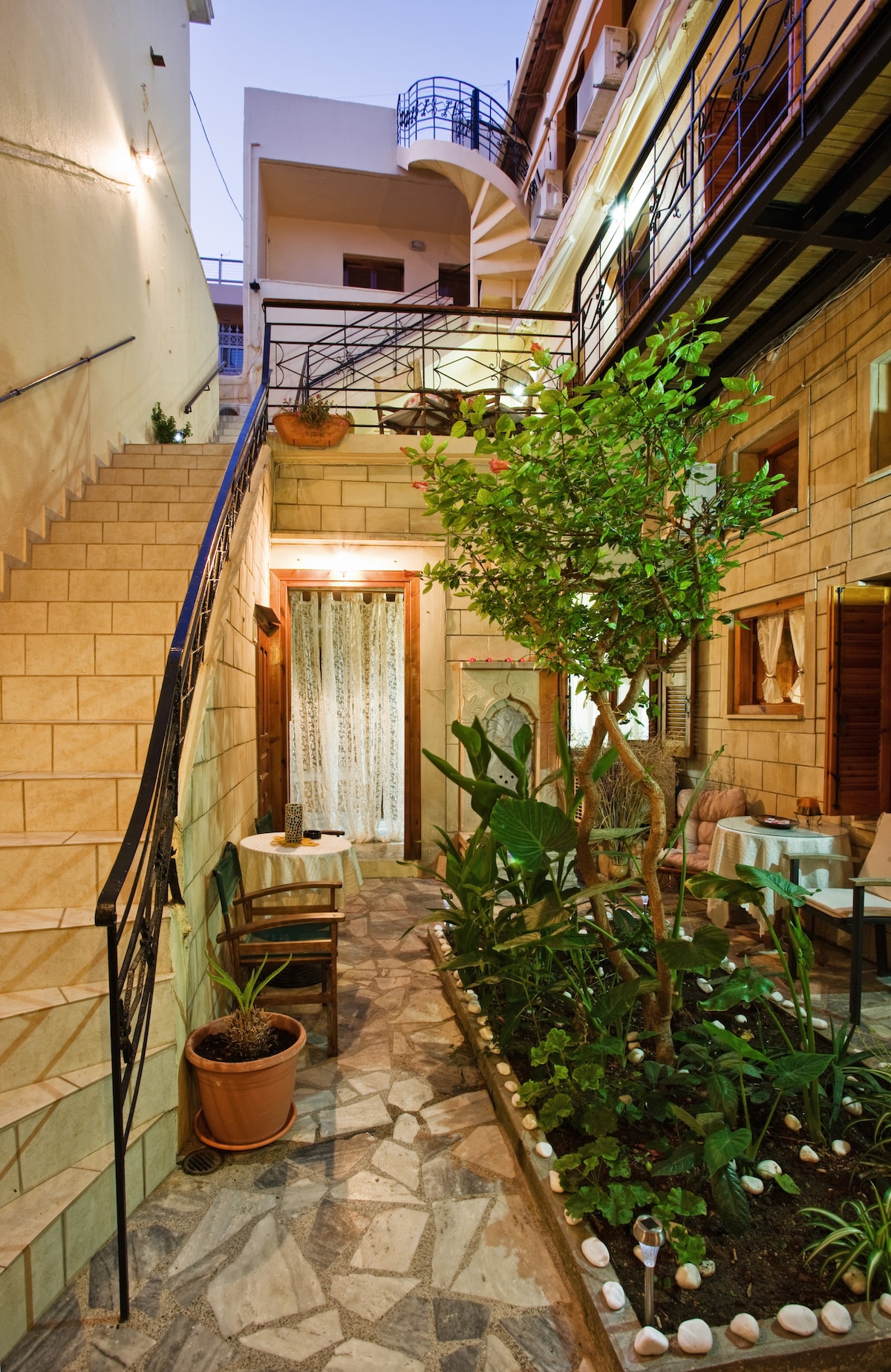 A charming courtyard is framed by stone walls, showcasing a pathway lined with greenery and decorative stones. Tables are placed under soft lighting, with stairs leading to upper levels. A light breeze can be felt in this inviting communal space, adding to the tranquil ambiance.