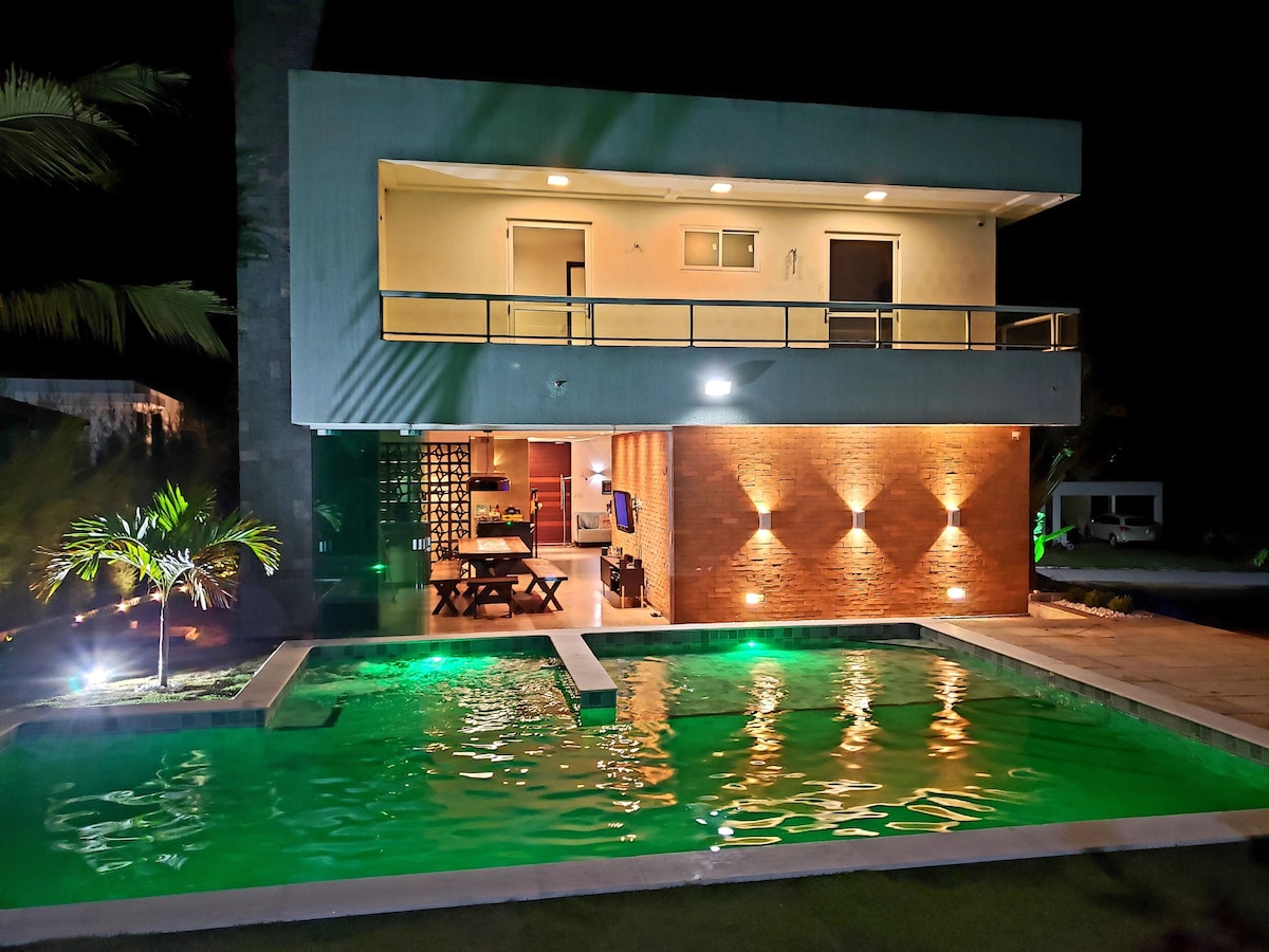 A modern two-story home is highlighted by a well-lit exterior and a tranquil pool reflecting green hues. The open patio features a dining area, while large windows allow visibility into the inviting interior space.