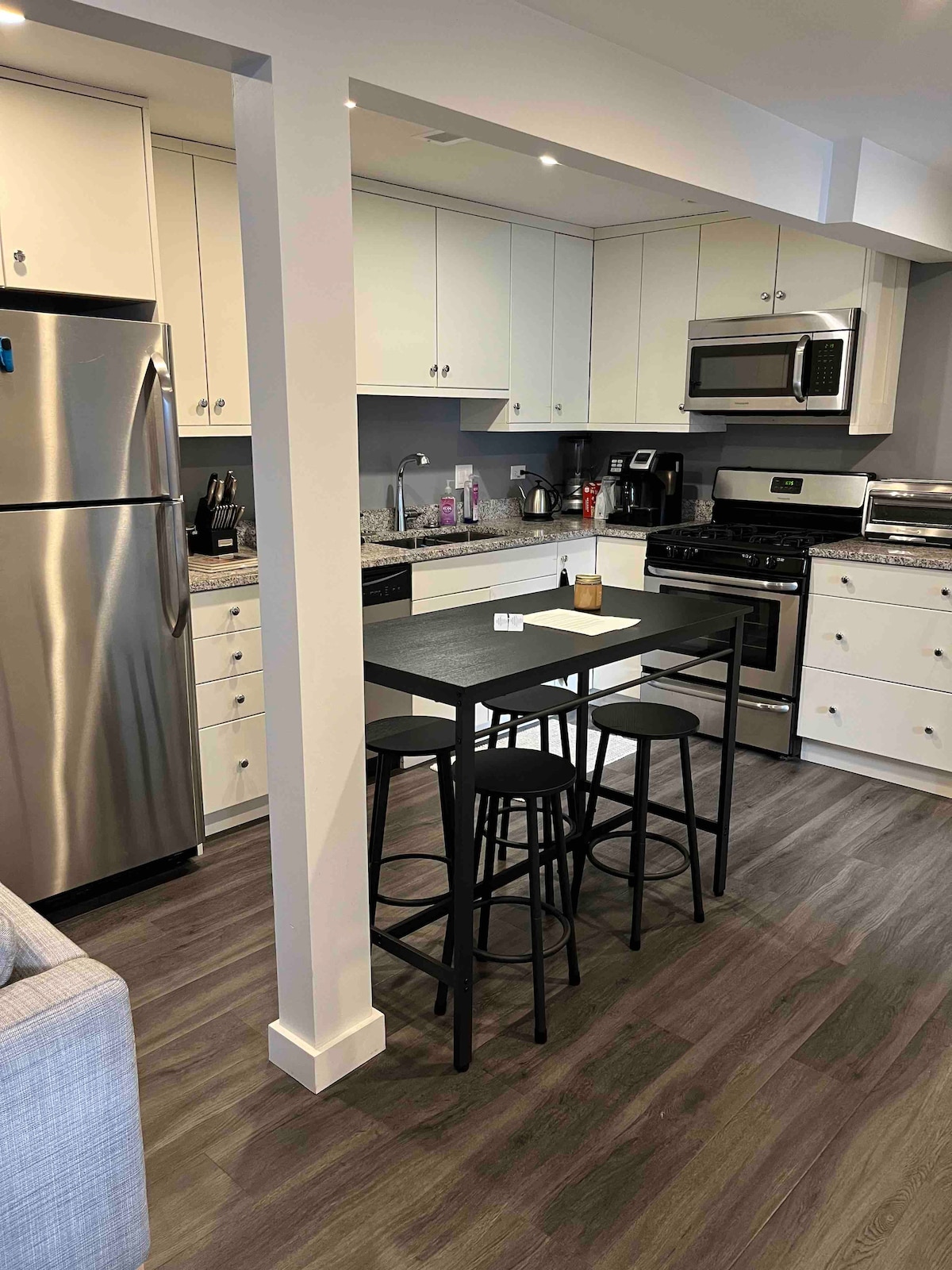 The kitchen features modern, streamlined cabinetry and a stainless steel refrigerator. A small dining table with three black stools offers a casual dining space. The kitchen’s layout includes a stove, microwave, and sink, with counter space for meal preparation.