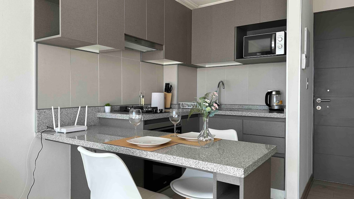 The modern kitchen area is equipped with sleek cabinets and a spacious countertop, featuring essential appliances including a microwave and coffee maker. A small dining table is set with two plates and glasses, complemented by a decorative vase. Natural light brightens the space.