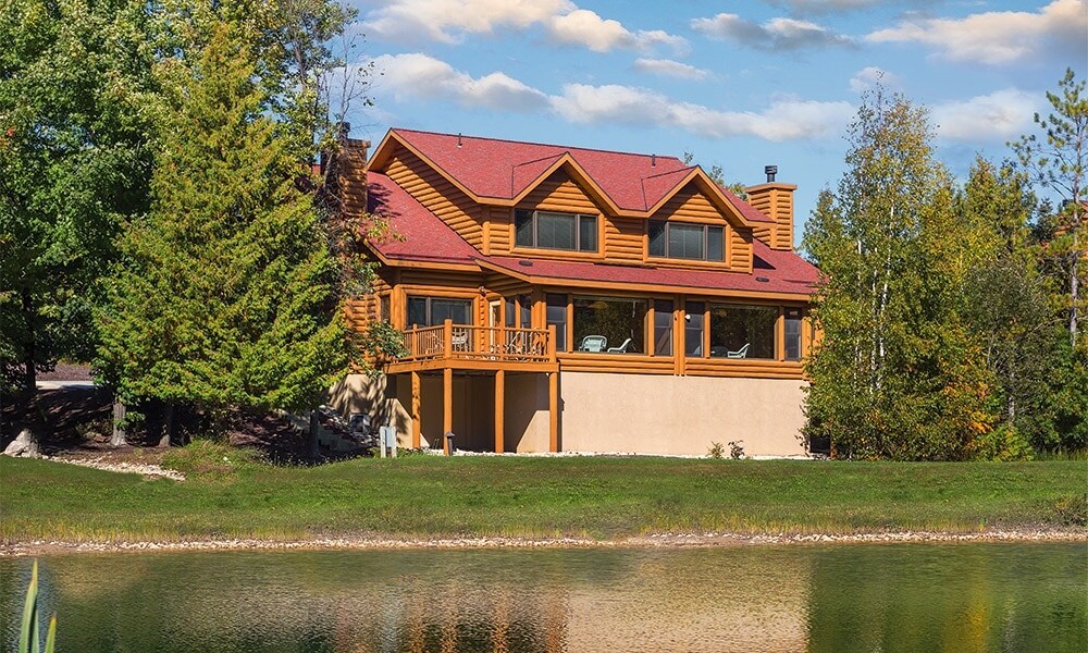 The exterior of a large log cabin is shown, featuring a red roof and multiple large windows. The structure overlooks a tranquil pond, surrounded by lush green trees. A balcony extends from the cabin, providing outdoor seating and views of the serene landscape.