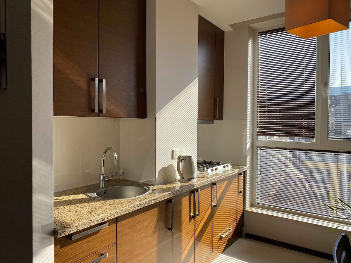 A modern kitchen is displayed, featuring sleek wooden cabinetry with metallic handles. A stainless steel sink is positioned next to a kettle. Natural light filters through a large window with horizontal blinds, illuminating the granite countertop and cooking range.
