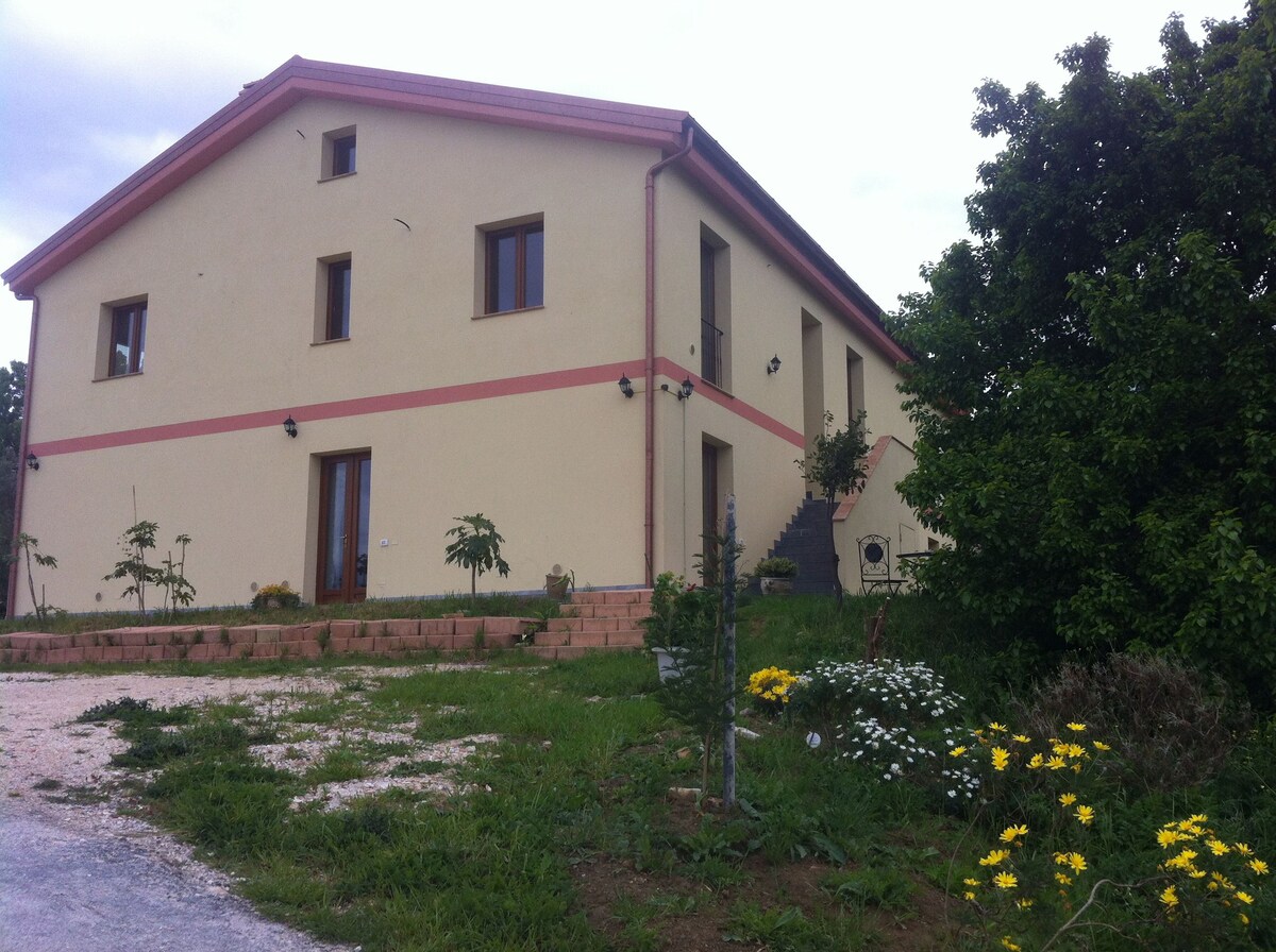The agriturismo building features a two-story structure with a soft beige exterior and pink accents. Surrounding gardens boast various plants and flowers, contributing to a serene rural atmosphere. Several windows allow natural light, and a pathway leads towards the entrance.
