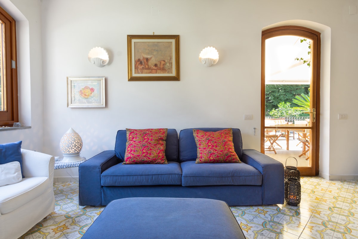 A living area is showcased with a blue sofa adorned with colorful cushions. Two light fixtures are mounted on the walls, and a framed artwork is displayed above the sofa. Natural light filters through large windows, illuminating the patterned floor tiles.