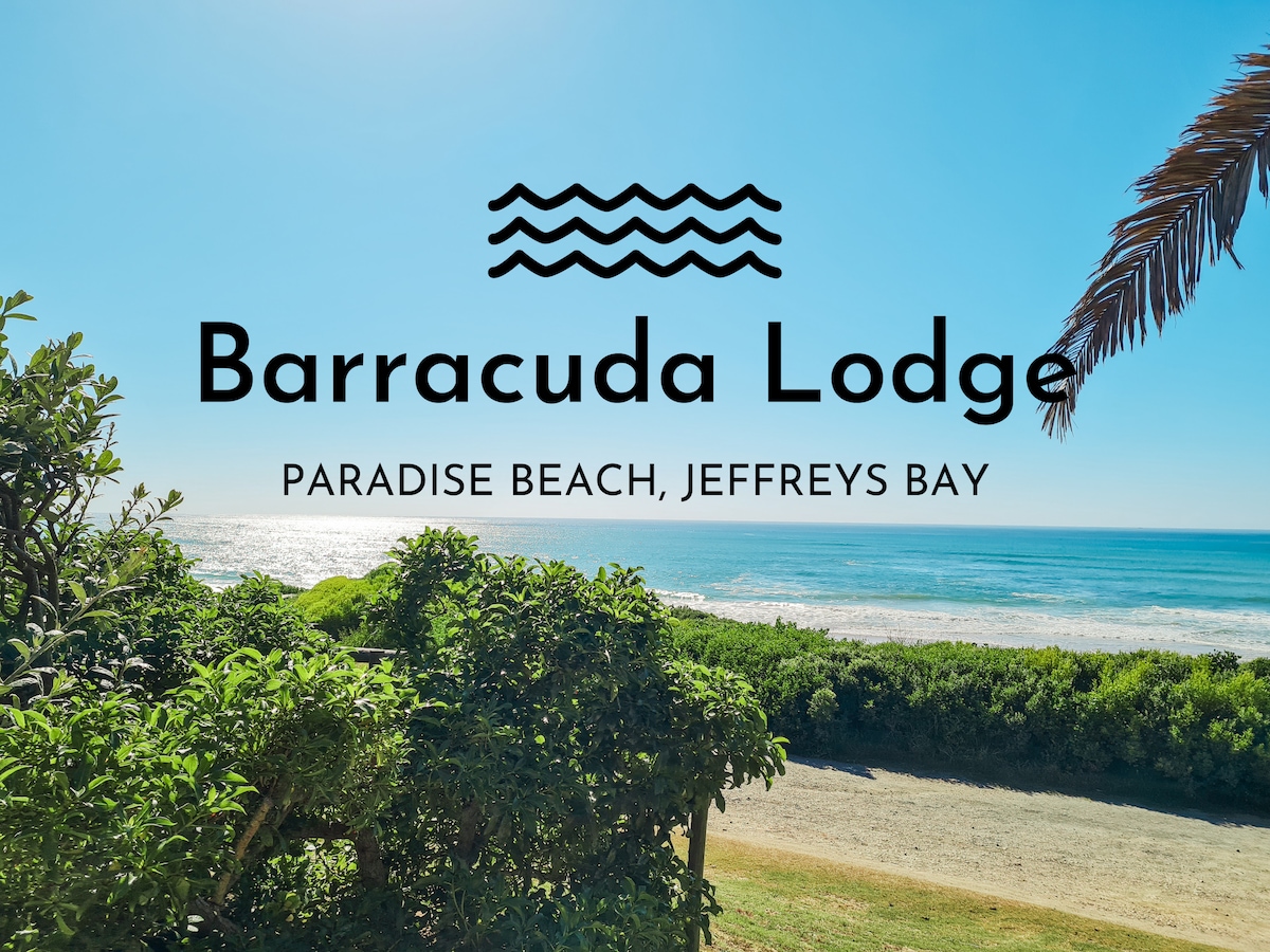 A vibrant view of the ocean stretches across the horizon, framed by lush green vegetation. The bright blue sky complements the inviting sea, while the serene landscape creates a peaceful beach atmosphere. The name 'Barracuda Lodge' is displayed prominently in elegant lettering.