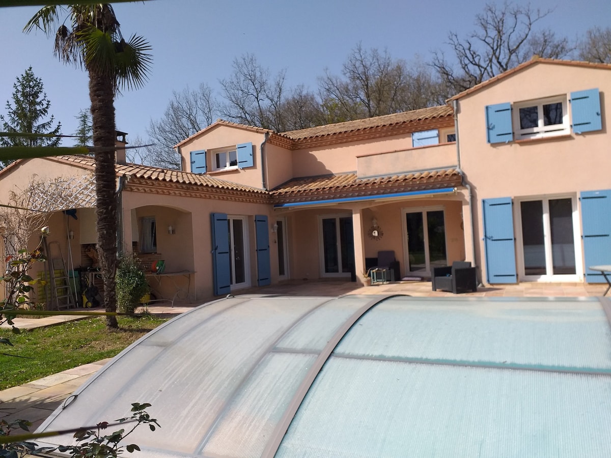 The exterior of a two-story pink house is highlighted by blue shutters and a tiled roof. A covered outdoor sitting area is visible, as well as a palm tree in the foreground. The surrounding landscape includes greenery, suggesting a peaceful environment.
