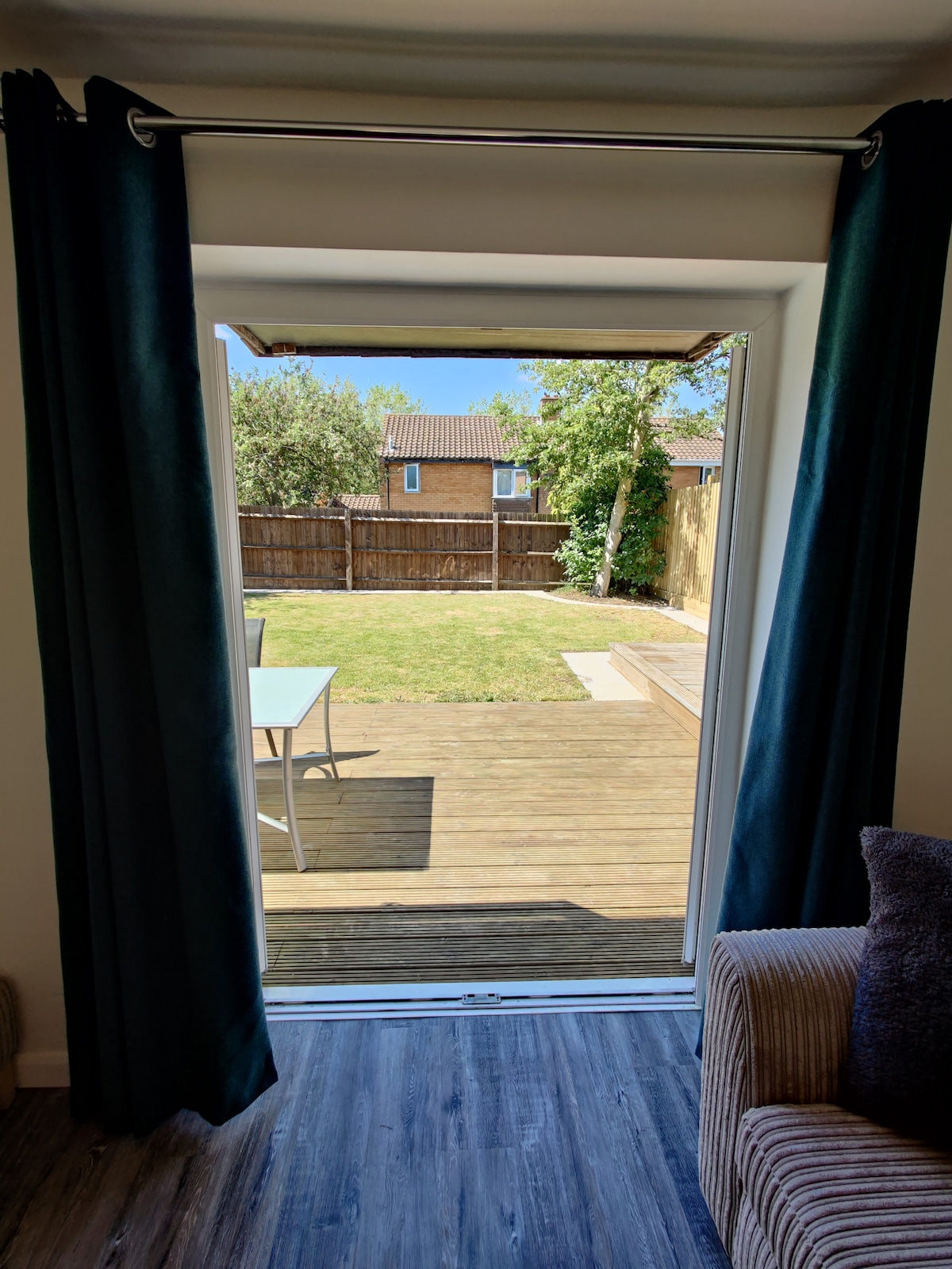 A view from inside the house showcases a sliding door leading to a spacious wooden deck and a grassy backyard. Sunlight fills the outdoor area, framed by green curtains and a soft, textured sofa visible in the foreground.